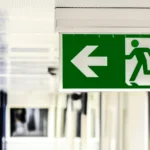A green emergency exit sign with a running figure and a left-pointing arrow hangs from the ceiling in a hallway.