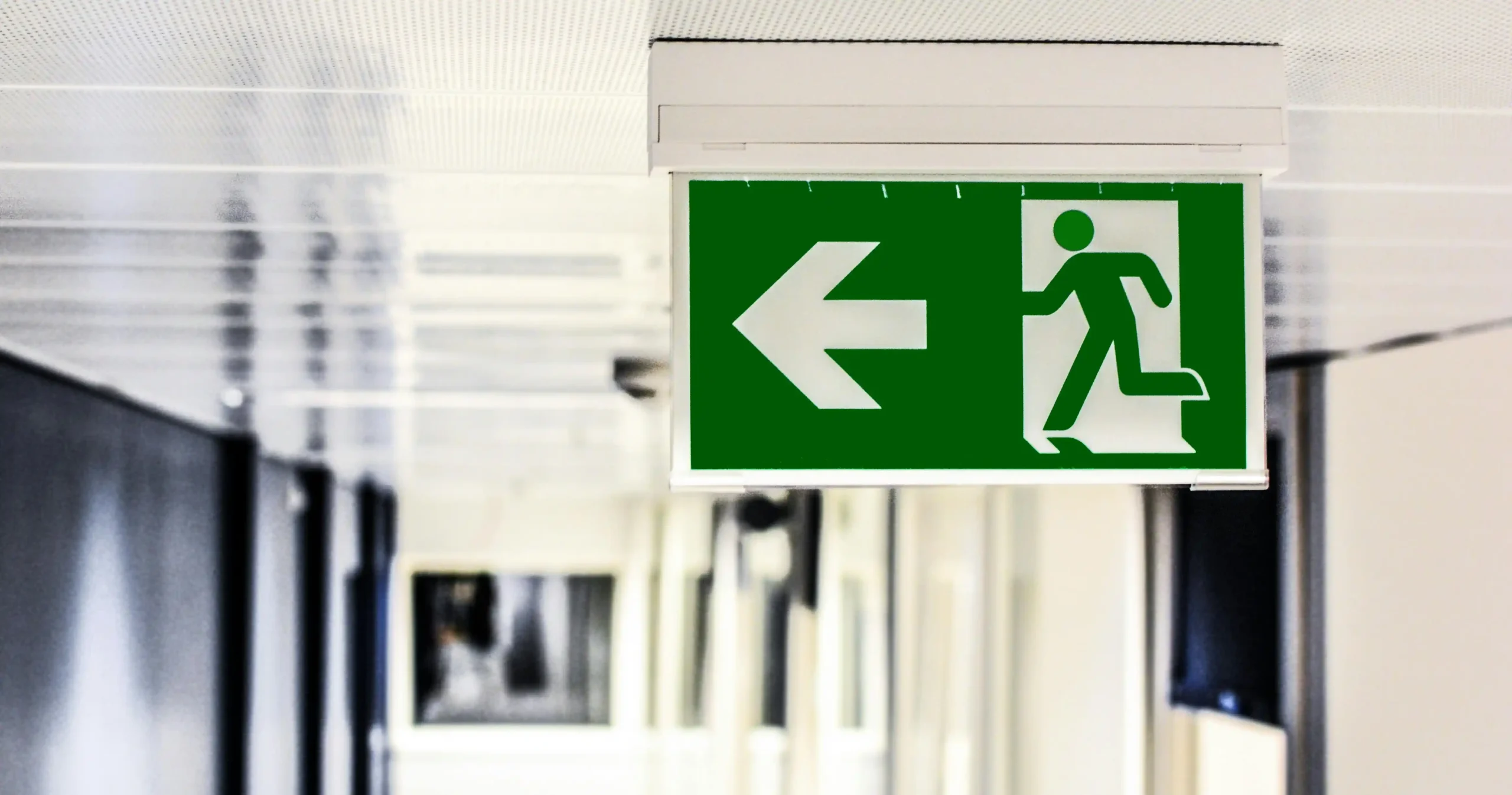 A green emergency exit sign with a running figure and a left-pointing arrow hangs from the ceiling in a hallway.